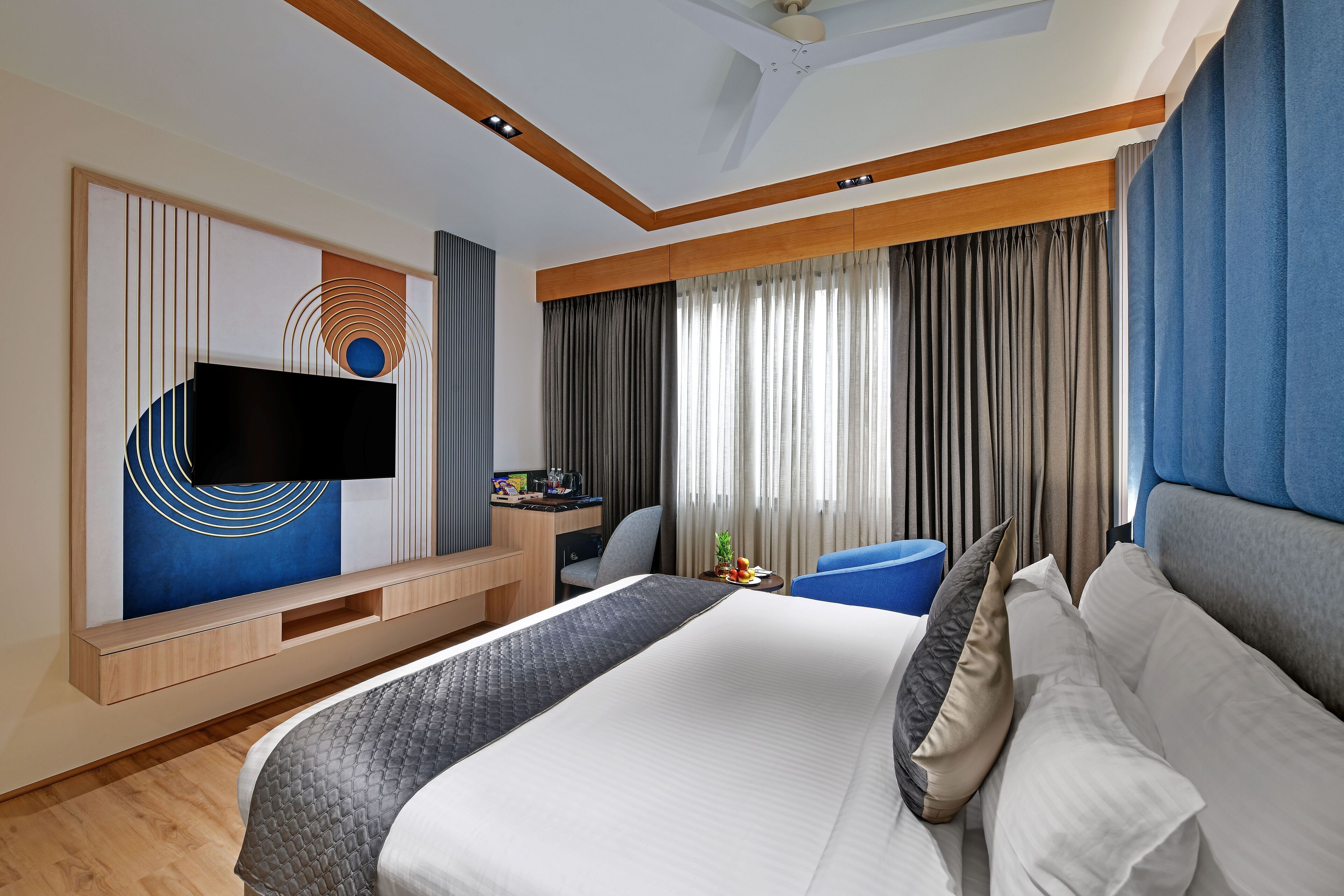 A modern hotel room with a large bed, a flat-screen TV mounted on the wall, a desk with a chair, and a large window with curtains.