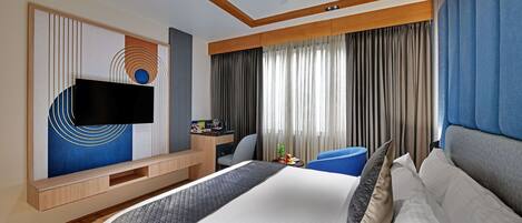 A modern hotel room with a large bed, a flat-screen TV mounted on the wall, a desk with a chair, and a large window with curtains.