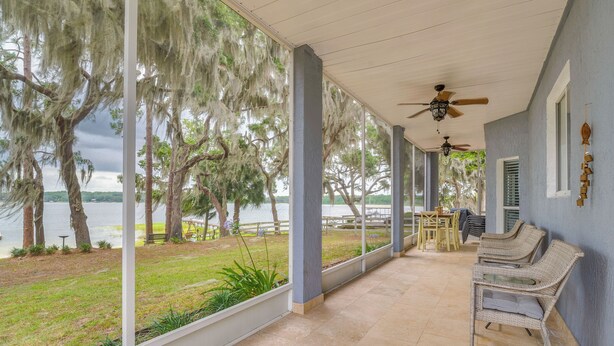 A covered porch with a view of a lake and trees, featuring wicker chairs and a ceiling fan. - Private Beach at Lakefront Summerfield Getaway! (Summerfield)