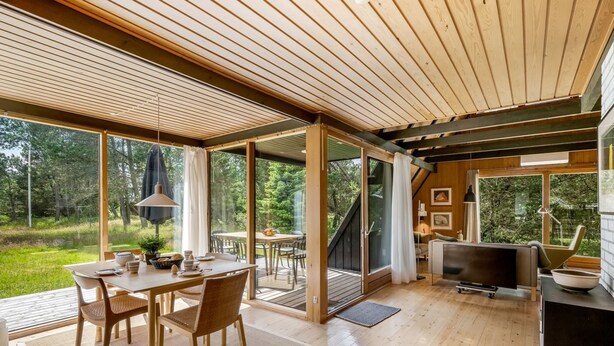 Un interior moderno con piso de madera, un área de comedor con sillas y una mesa, un sofá, un televisor y ventanas amplias que ofrecen vistas al exterior.