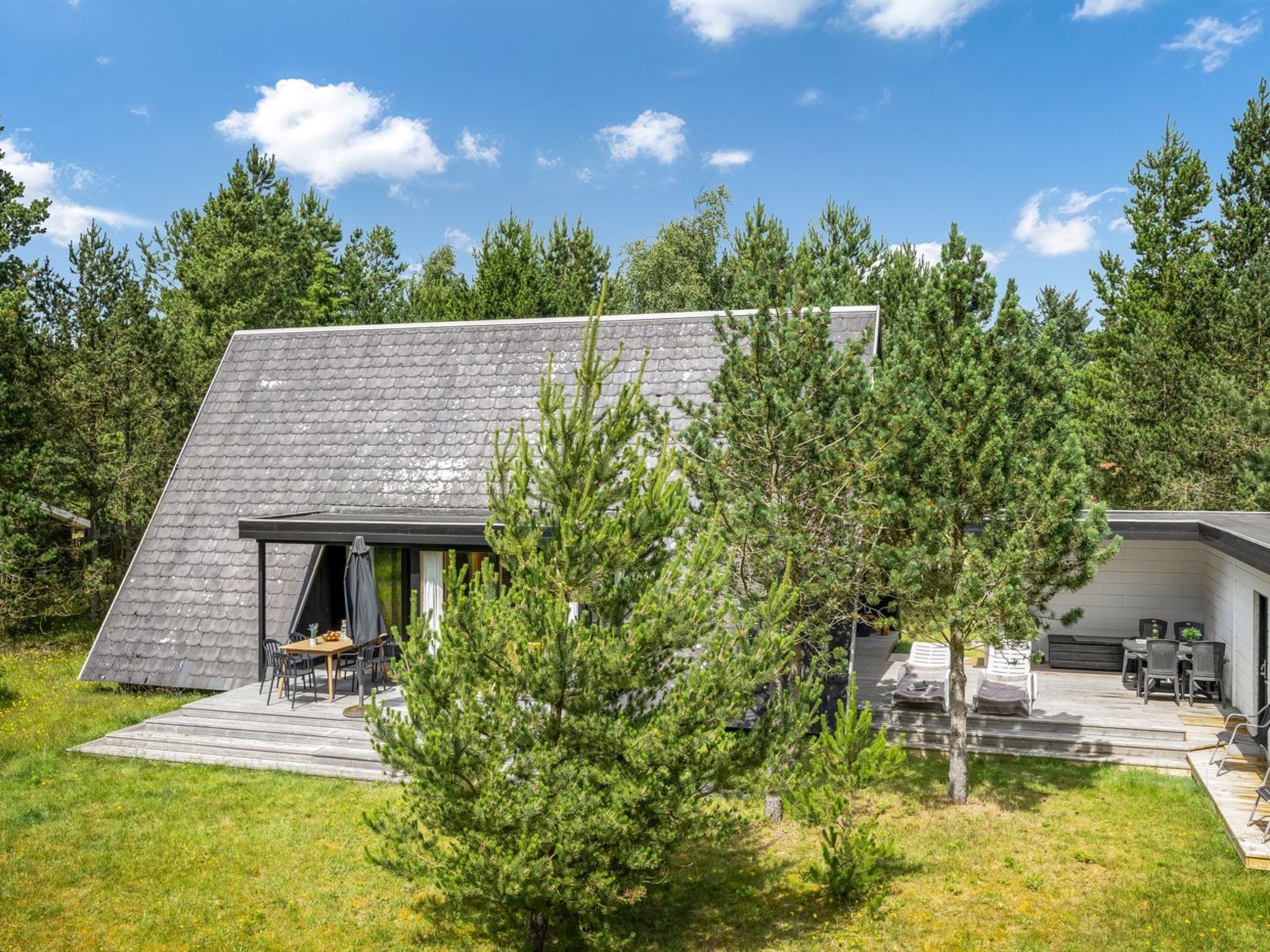 Une maison moderne avec un toit en pente, une terrasse dotée de mobilier d’extérieur et entourée d’une forêt.