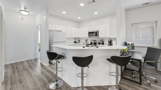A modern kitchen with white cabinets, a central island with bar stools, and a dining area with a table and chairs. - Tranquil 3BR I Free Parking I Workspace (Grand Prairie)