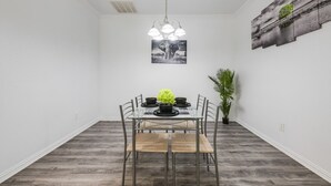 A dining area with a glass table, metal chairs, a black and white artwork above, and a potted plant on the right. - Tranquil 3BR I Free Parking I Workspace (Grand Prairie)