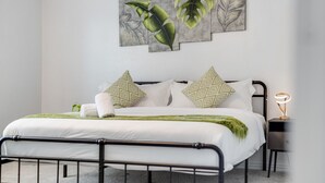 A modern bedroom with a bed, pillows, a nightstand, and a wall-mounted light. - Tranquil 3BR I Free Parking I Workspace (Grand Prairie)