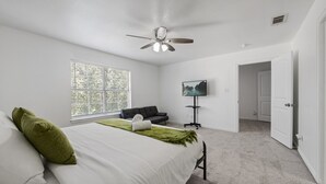 A bedroom with a bed, a black sofa, a TV on a stand, and a ceiling fan. - Tasteful 3BR I Free Parking I Workspace (Grand Prairie)