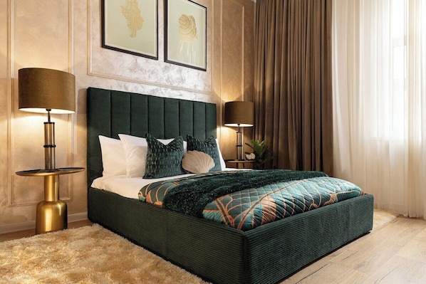 A large bed with a dark green upholstered headboard, multiple pillows, and a patterned bedspread. There are two bedside tables with lamps, a large window with curtains, and framed artwork on the wall.