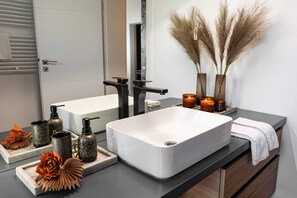 A modern bathroom with a large white sink, black faucet, and a mirror. There are decorative items like pampas grass, candles, and a tray with flowers.
