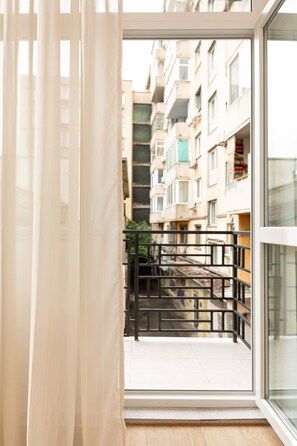 Blick durch ein Fenster mit Vorhang, der einen Balkon mit Metallgeländer und ein Gebäude im Hintergrund zeigt.