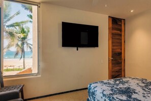 A bedroom with a bed, a television mounted on the wall, a window with a beach view, and a wooden door. - Your Private Piece of Mexican Beachfront Luxury (Playa Blanca)