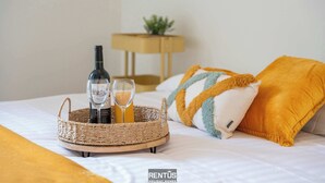 Una cama con una bandeja tejida que sostiene una botella y vasos, una pequeña planta en una mesita de noche y un almohadón amarillo.