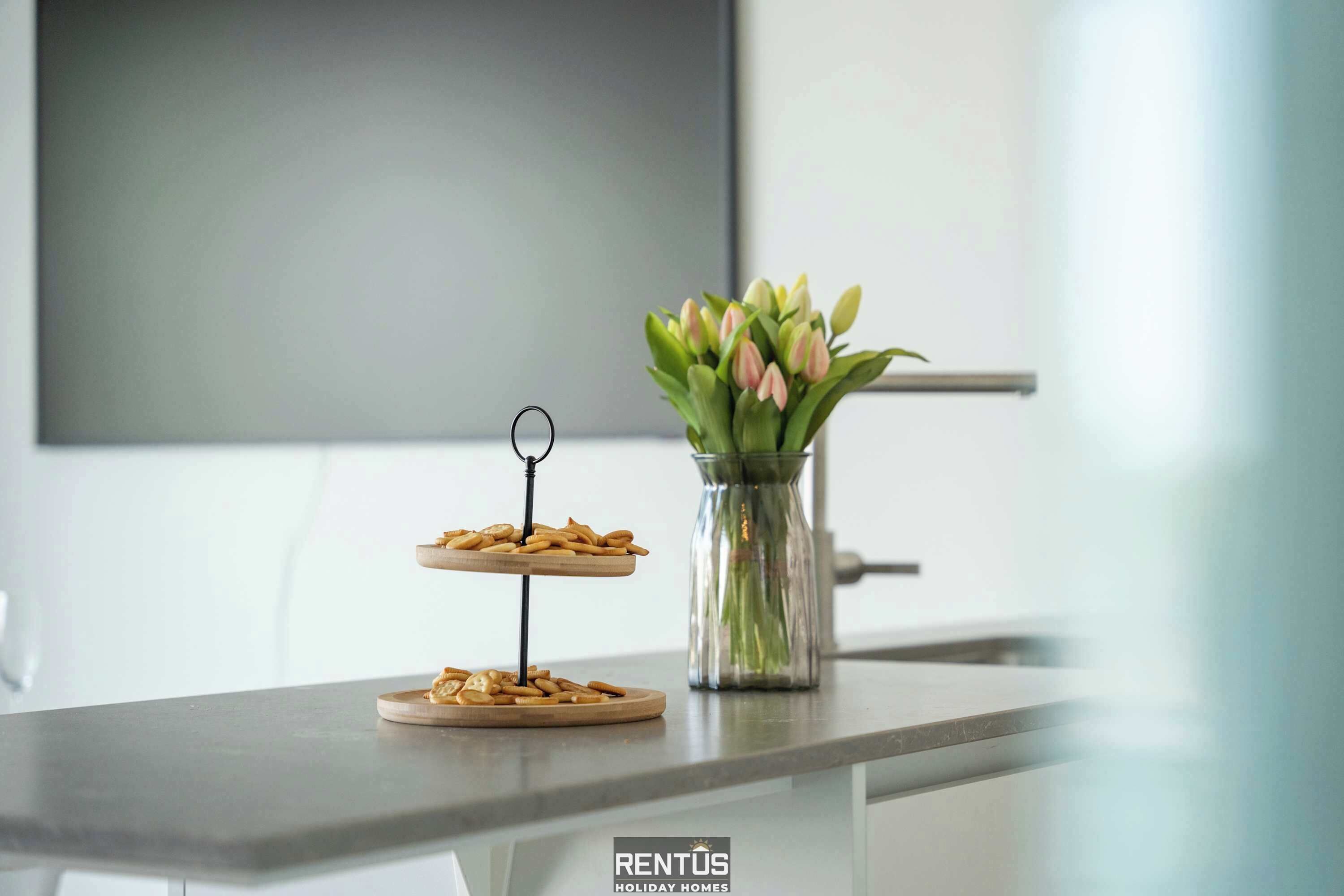 A kitchen counter with two tiered serving dishes of snacks, a vase with flowers, and a flat-screen TV in the background.