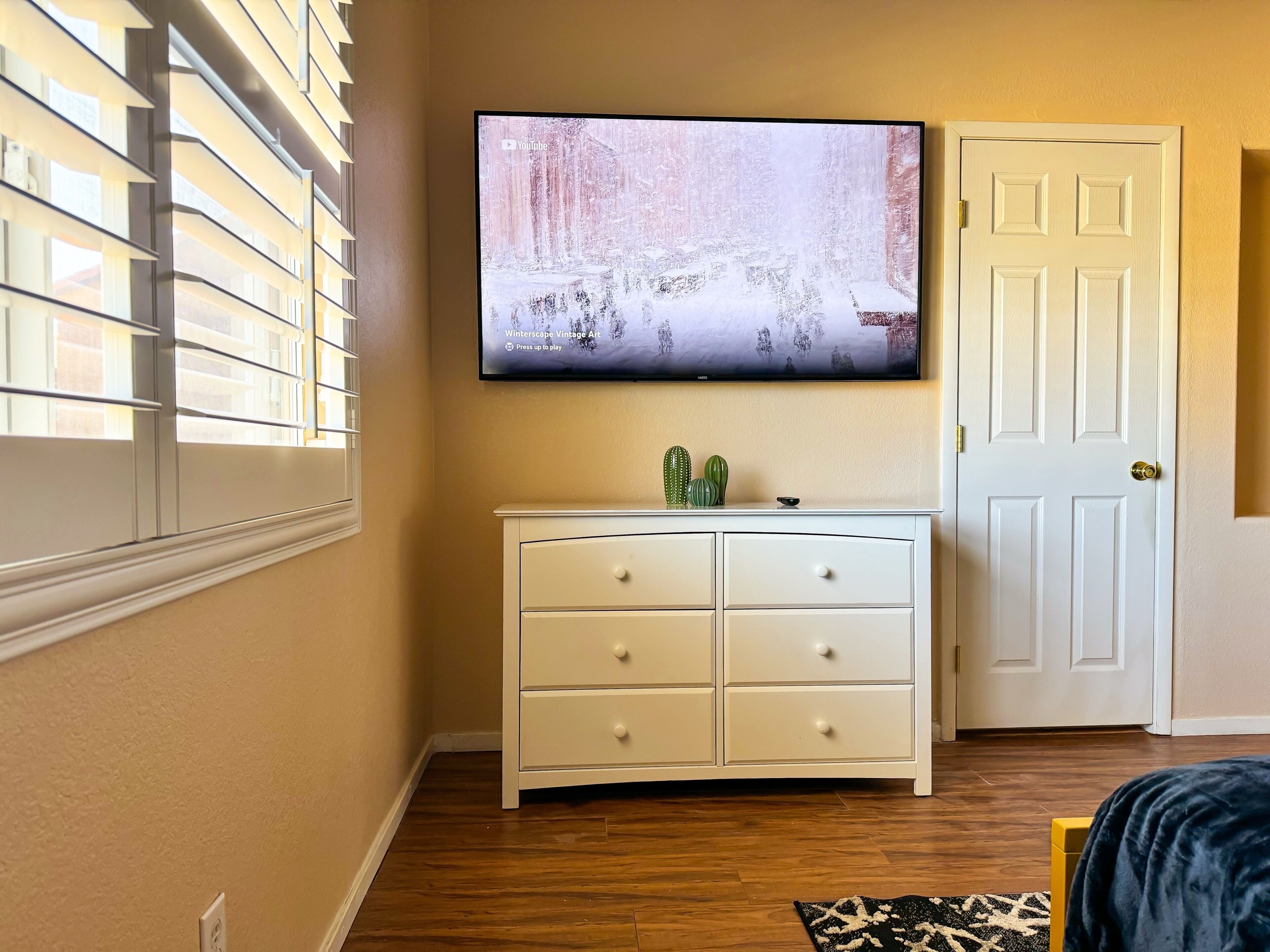 Een kamer met een tv aan de muur die een winterlandschap toont, een witte dressoir met laden, een deur en een raam met jaloezieën.