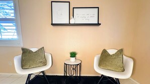 Una habitación con dos sillas blancas, un tapete negro, una mesita con una planta y dos cuadros enmarcados en la pared.