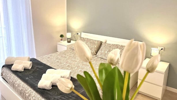 A neatly made bed with rolled towels and a bouquet of white tulips in a modern bedroom with a nightstand and bedside lamp. - Archivio 32 Neapolitan Apartment (Napoli)
