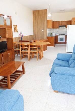 A modern living room with a wooden dining table, blue sofa, and a kitchen area with wooden cabinets and a white refrigerator. - Kritos Apartments with Two Bedrooms (Argaka)