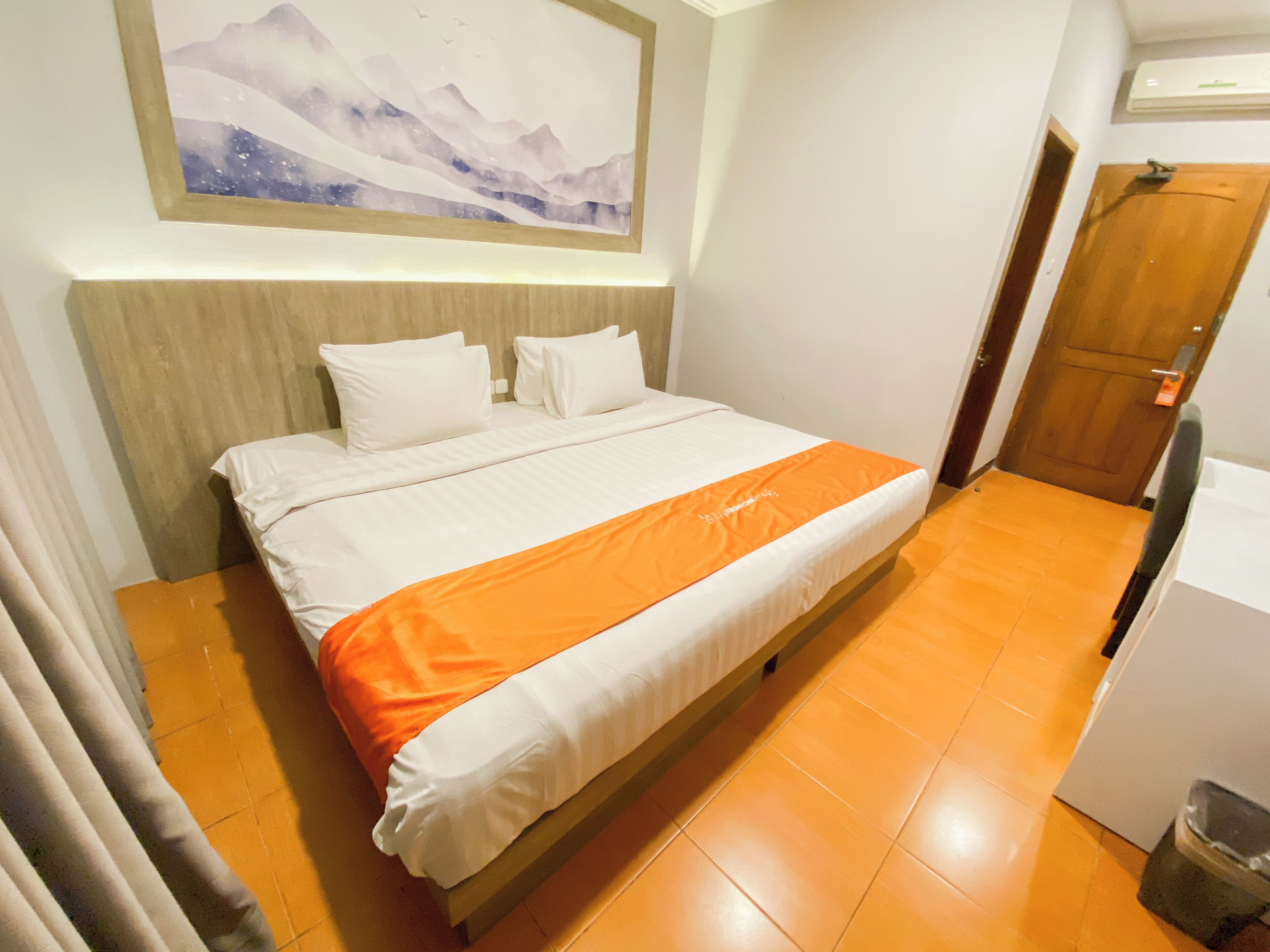 A hotel room with a bed, a wooden headboard, an orange bedspread, and a painting on the wall.