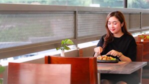 Une personne est assise à une table devant un repas, dans une pièce dont la fenêtre, ornée d’une grille métallique, offre une vue sur l’extérieur.