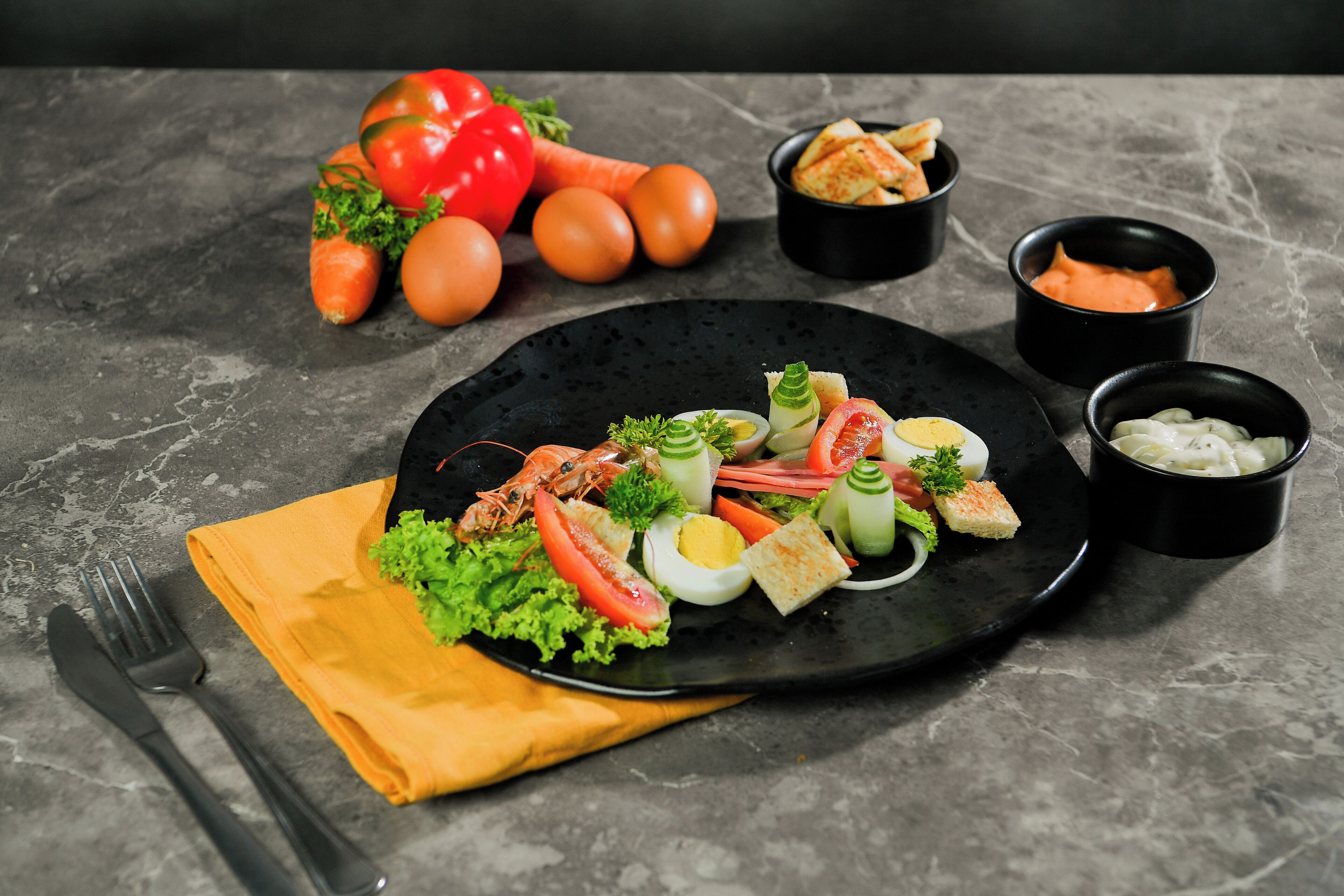 A black plate with a variety of food items including vegetables, eggs, and bread, accompanied by a fork and knife on a napkin.