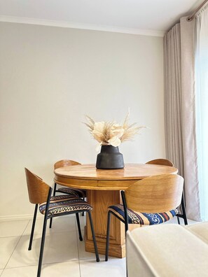 Une table de salle à manger ronde en bois, sur laquelle est posé un vase noir contenant des fleurs séchées, entourée de quatre chaises dotées de coussins à motifs.