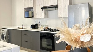 A modern kitchen with grey cabinets, a stainless steel refrigerator, a stove, and a washing machine. - The Oasis 92 on New Midrand (Midrand)
