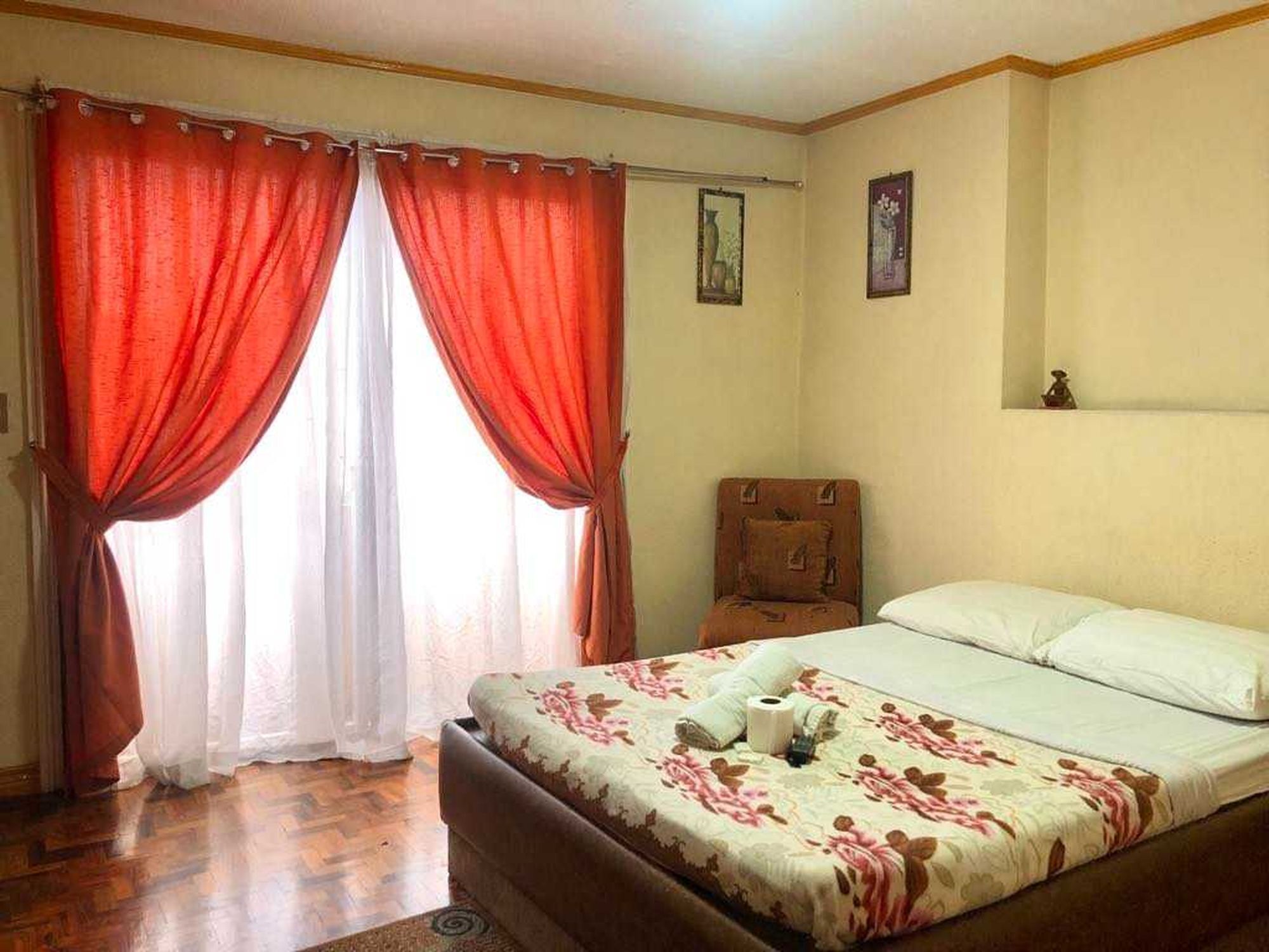 A bedroom with a bed, a chair, a window with red curtains, and a painting on the wall.
