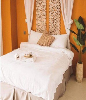 Una cama bien tendida con sábanas blancas, un cojín estampado y una mesita de noche con un libro y una caja de pañuelos.