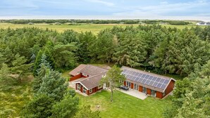 Une maison à deux étages, à l’extérieur rouge et dotée d’un toit recouvert de panneaux solaires, entourée d’une forêt dense et offrant une vue sur l’océan.