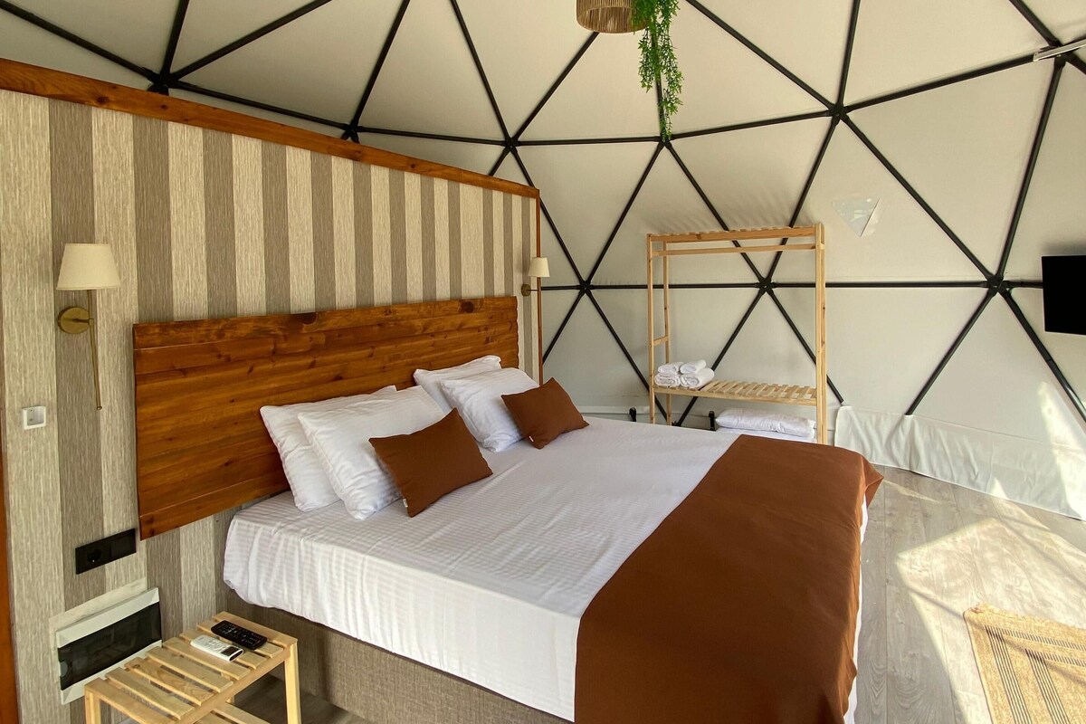 Quarto moderno com design geométrico no teto, cabeceira de madeira, cama com roupa de cama branca e marrom, mesa de cabeceira com abajur e planta pendente.