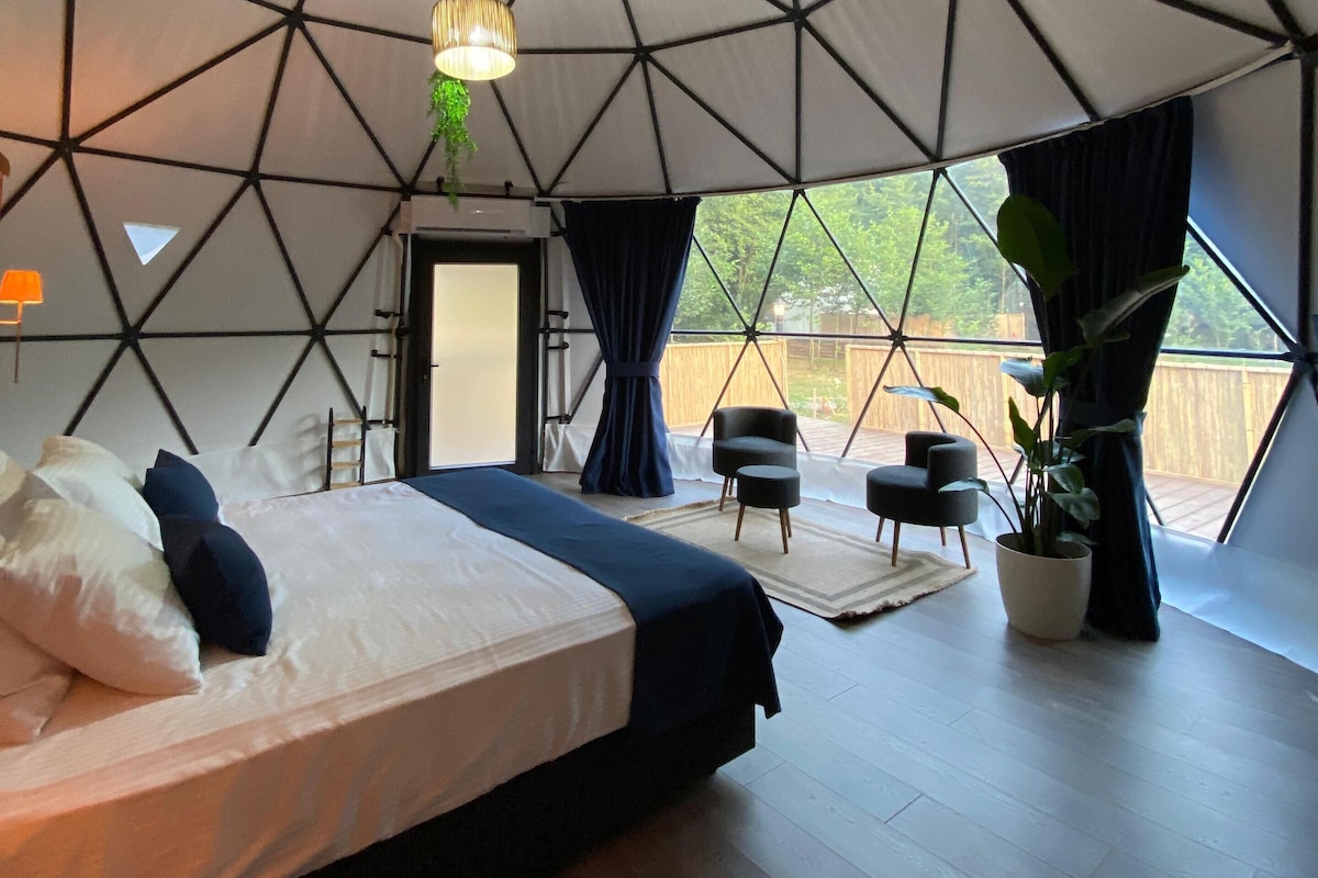 Quarto tipo tenda com cama, luminária pendente, planta em vaso e vista para o exterior.