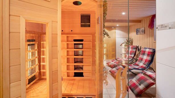 A sauna room with wooden benches and a glass door leading to a seating area with red and grey checkered cushions.
