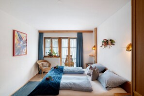A bedroom with a bed, a chair, a window with curtains, a wall-mounted light, and a painting.