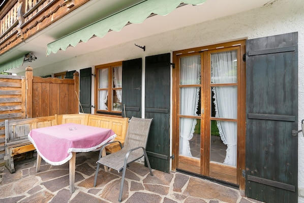 Un patio con suelo de piedra, un banco y una mesa de madera, una silla y una puerta con cortinas.