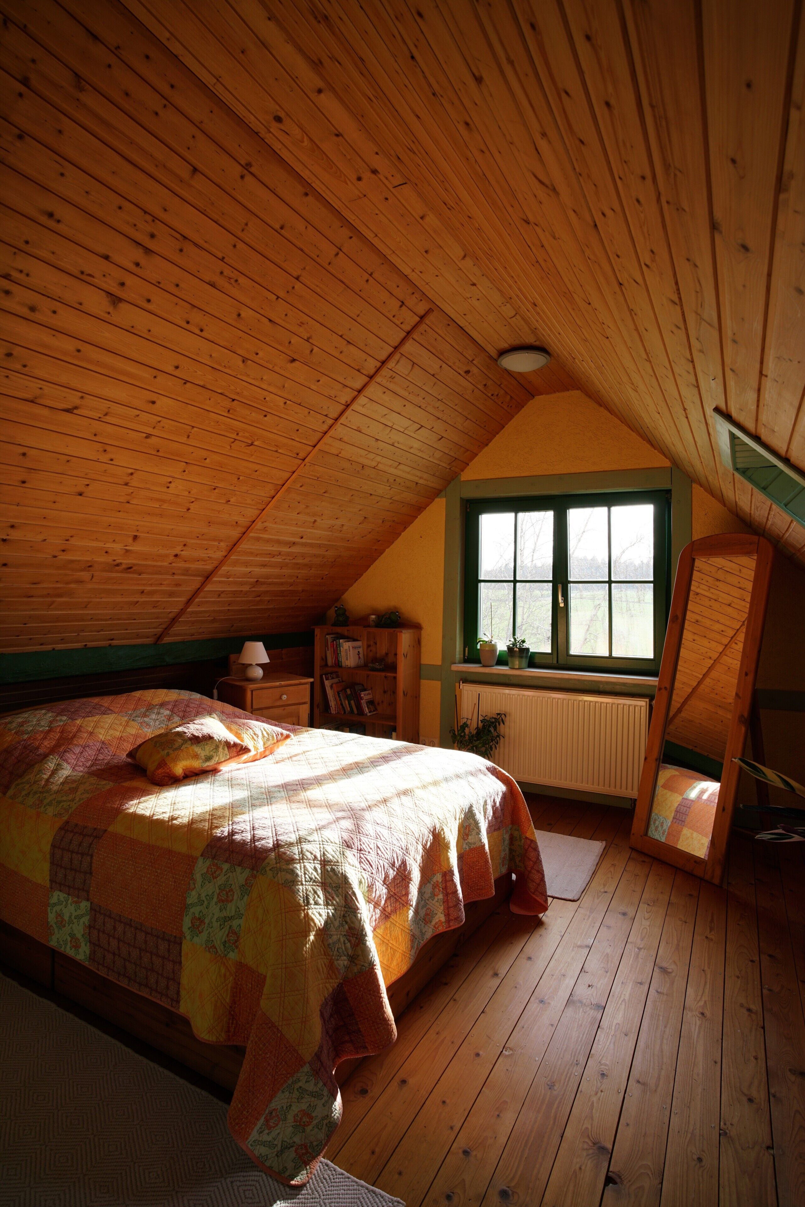 A bedroom with a sloped ceiling, a bed with a quilt, a window with shutters, a radiator, a nightstand, and a mirror leaning against the wall.