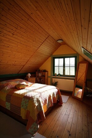 Ein Schlafzimmer mit geneigter Decke, einem Bett mit gesteppter Decke, einem Fenster mit Fensterläden, einem Heizkörper, einem Nachttisch und einem an der Wand angelehnten Spiegel.