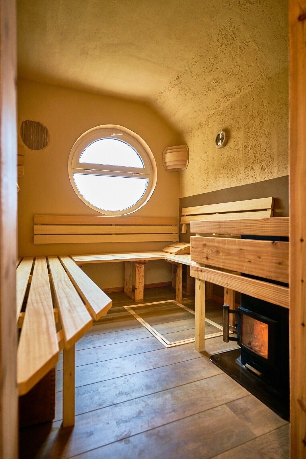Een sauna met houten banken, een rond raam en een houtkachel.