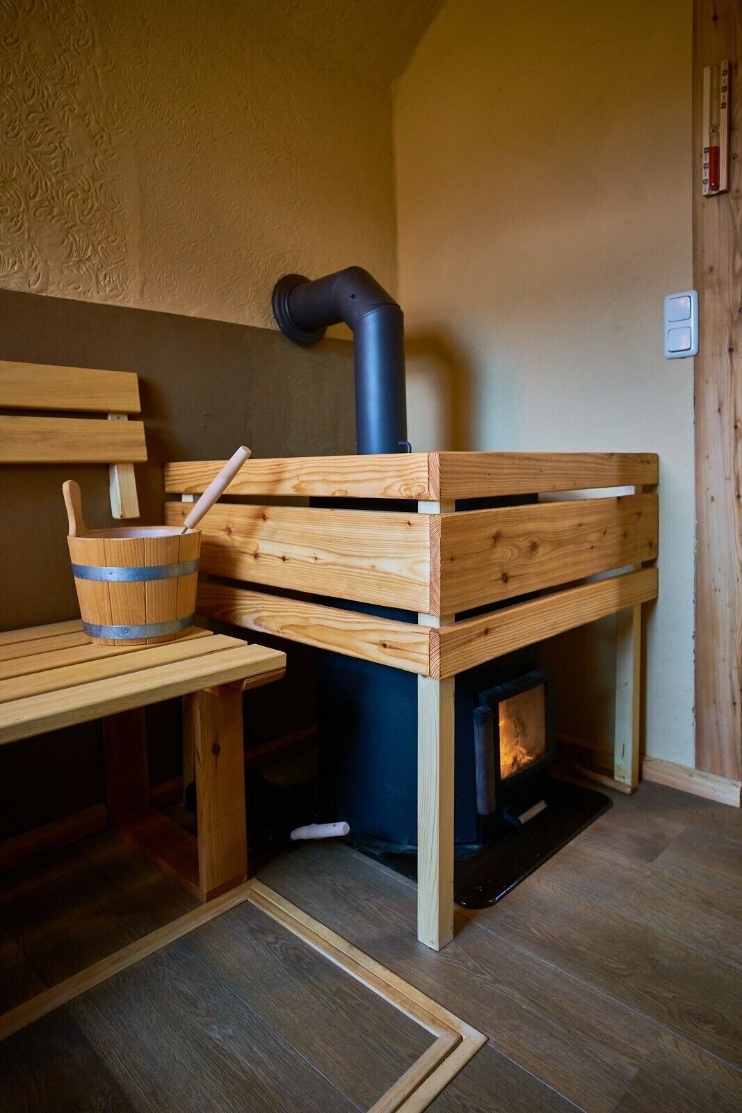 A sauna with wooden benches and a bucket, a black chimney, and a visible fire.