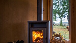 Un poêle à bois dont on peut voir les flammes, une poubelle noire et une fenêtre donnant sur des arbres.