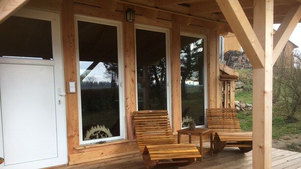 Une terrasse en bois avec deux chaises et une table en bois, une porte blanche et de grandes fenêtres.