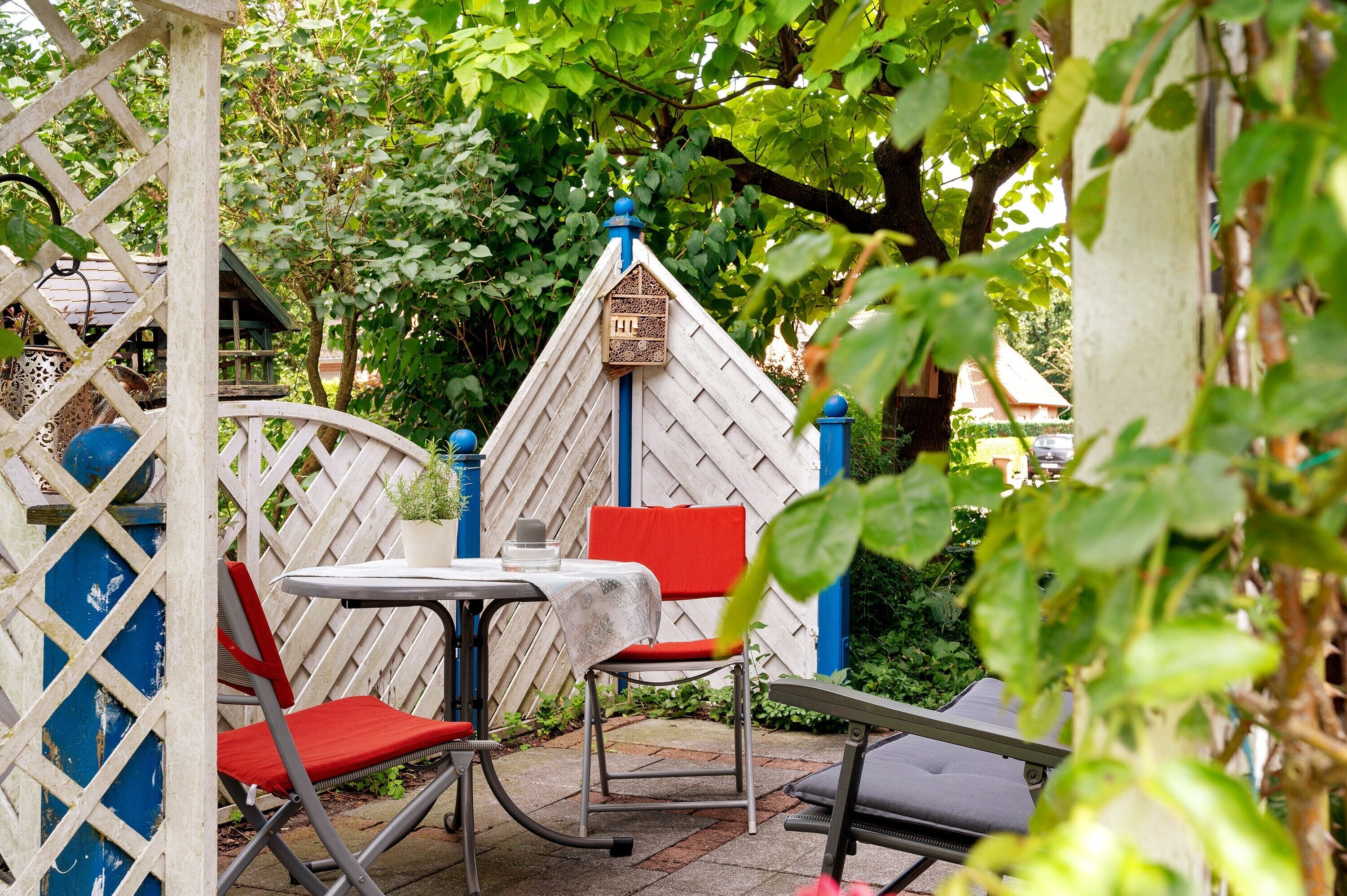 En patio med hvidt espalier, et lille bord med rød pude og en rød stol.