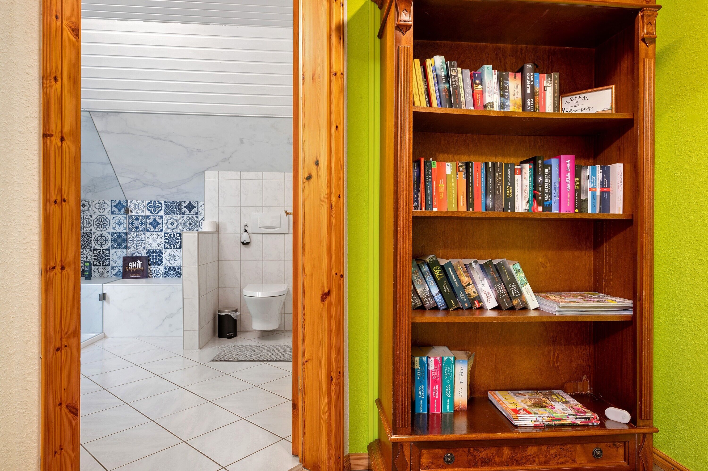 Casa de banho com sanita, lavatório e zona de duche. Uma estante de madeira com livros e revistas.