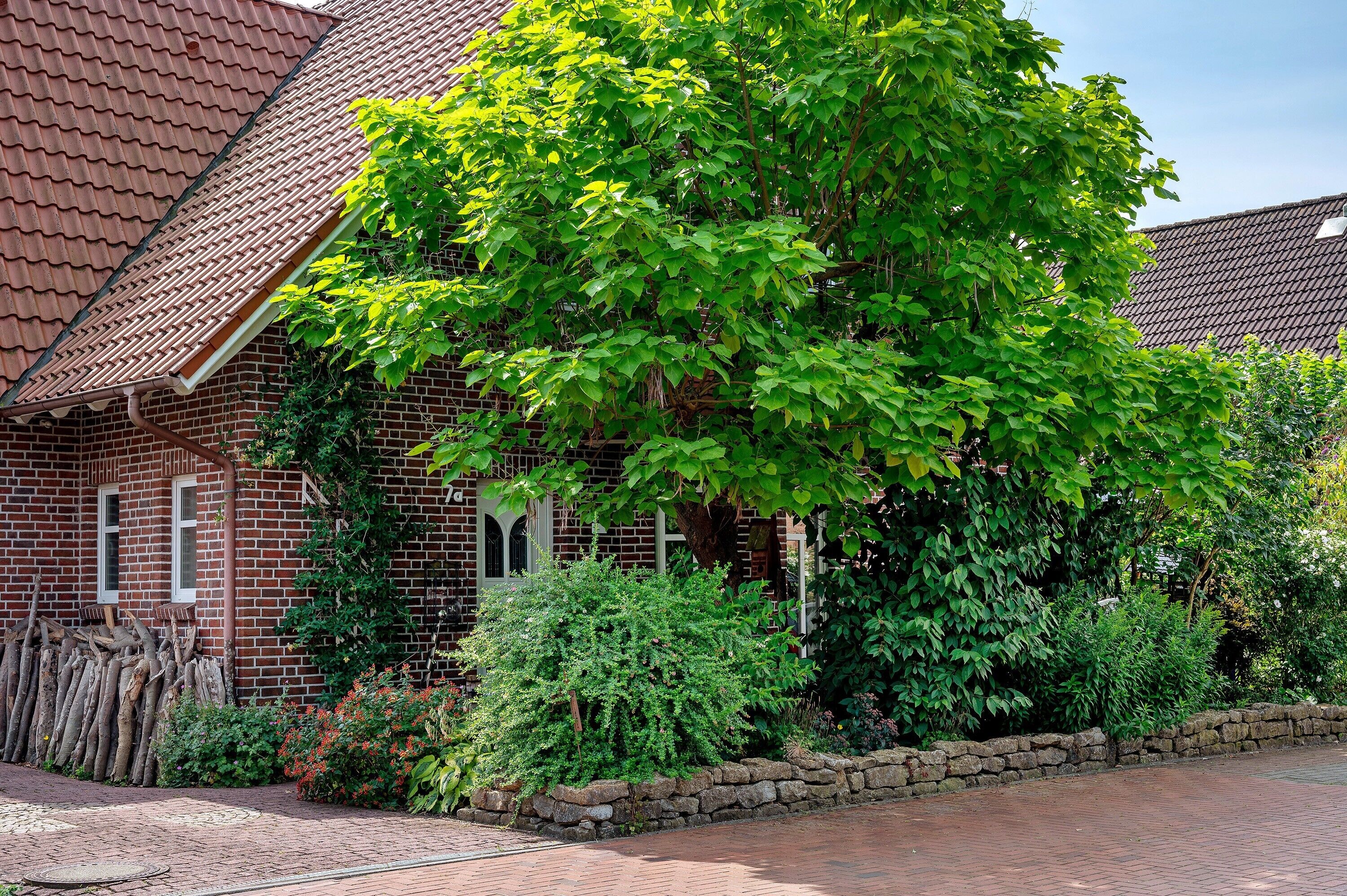 Ein Haus aus Backsteinen mit einem Ziegeldach, ein großer Baum davor und eine Steinmauer.