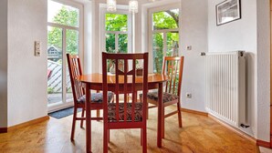 A dining area with a wooden table and chairs, large windows, a radiator, and a ceiling with wooden beams. - Apartment 'Inge Huus' with Private Terrace, Garden, and Wi-Fi (Hude)