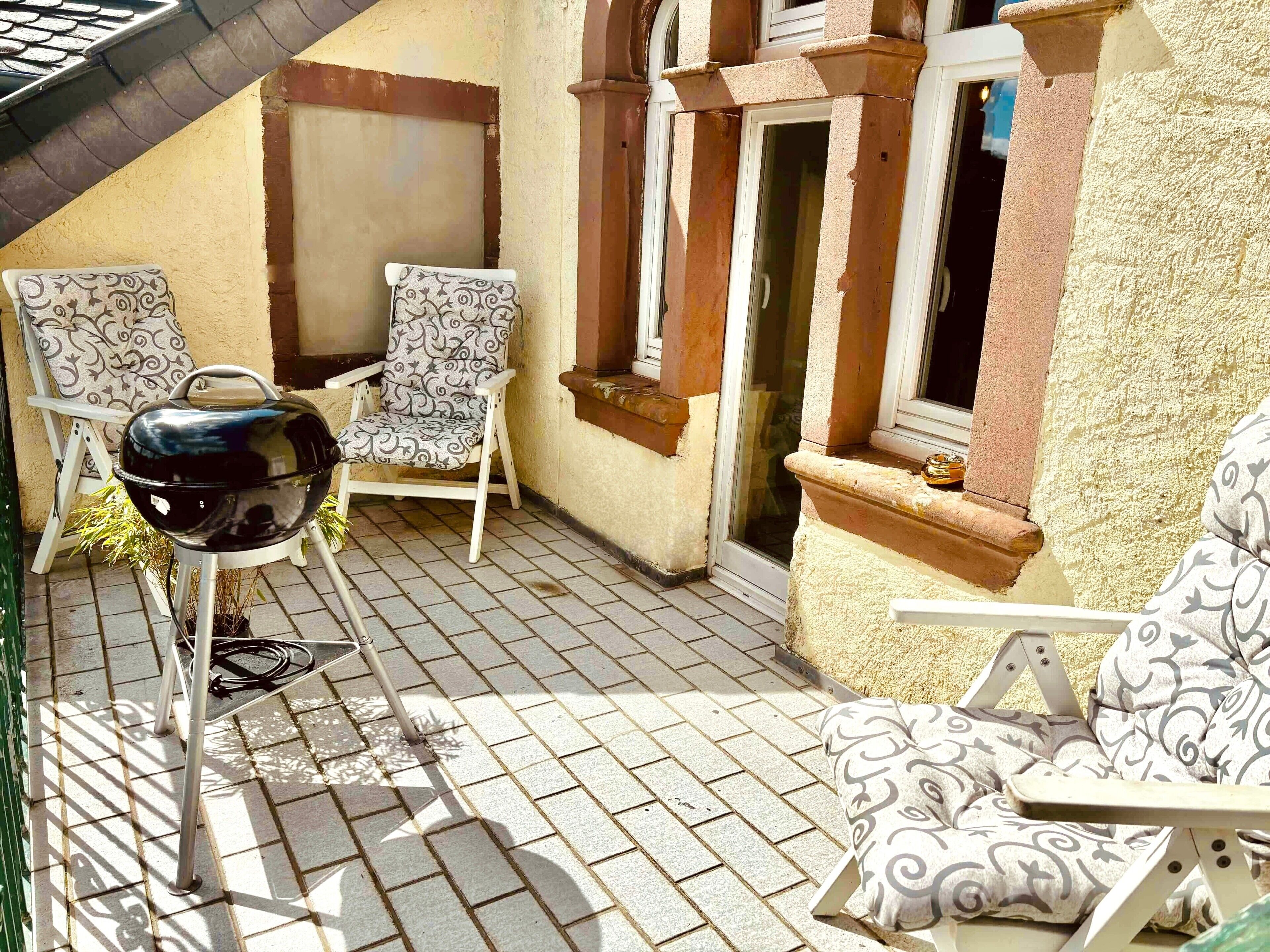 Une terrasse avec un barbecue, deux chaises à motifs et une fenêtre donnant sur un beau paysage.