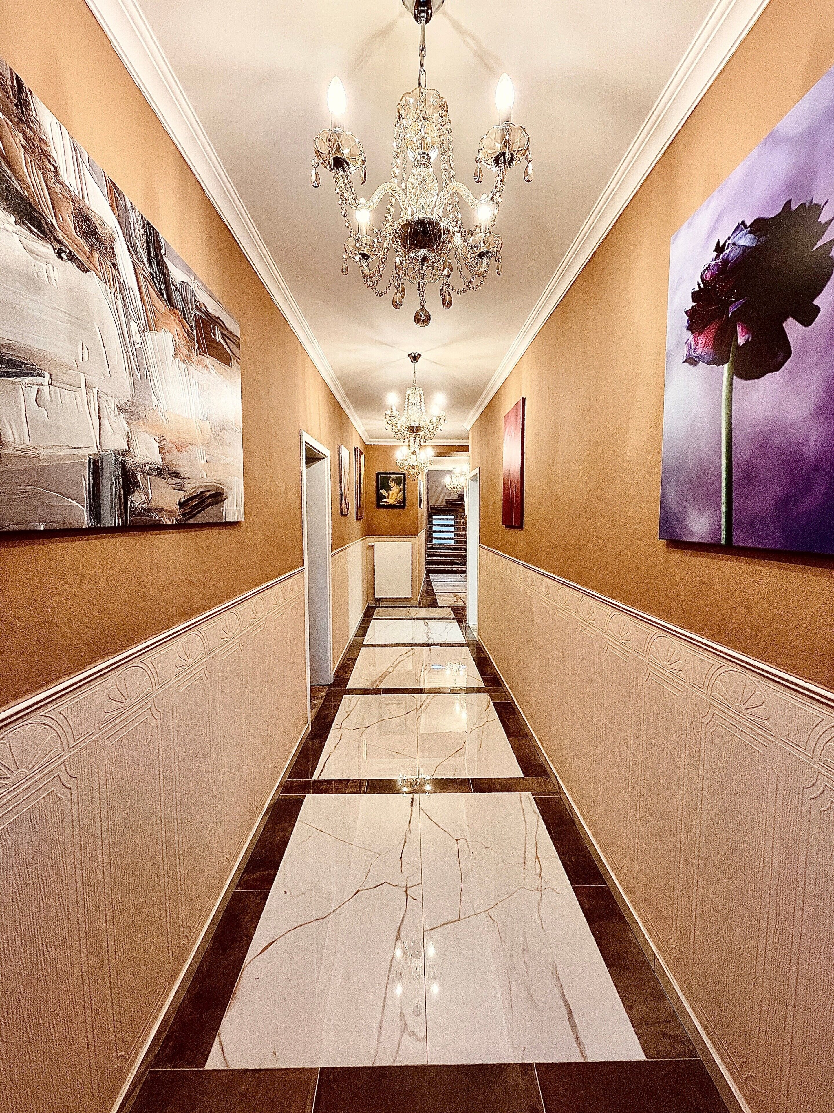 A hallway with a chandelier, marble flooring, and artwork on the walls.