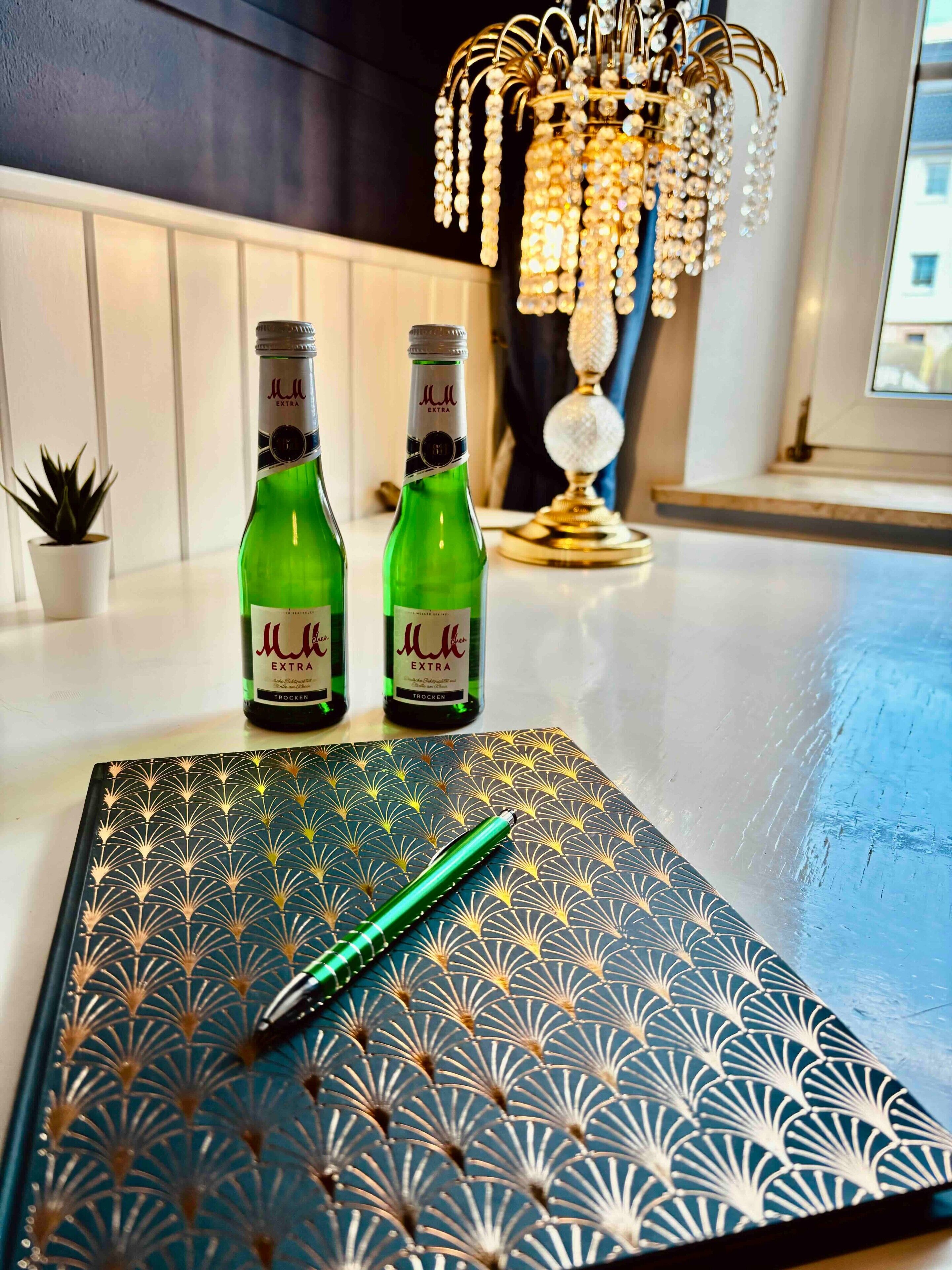 Deux bouteilles de « M.M. Extra » sont posées sur une table, accompagnées d’un stylo décoratif, d’un carnet à motifs et, en arrière-plan, d’un lustre.