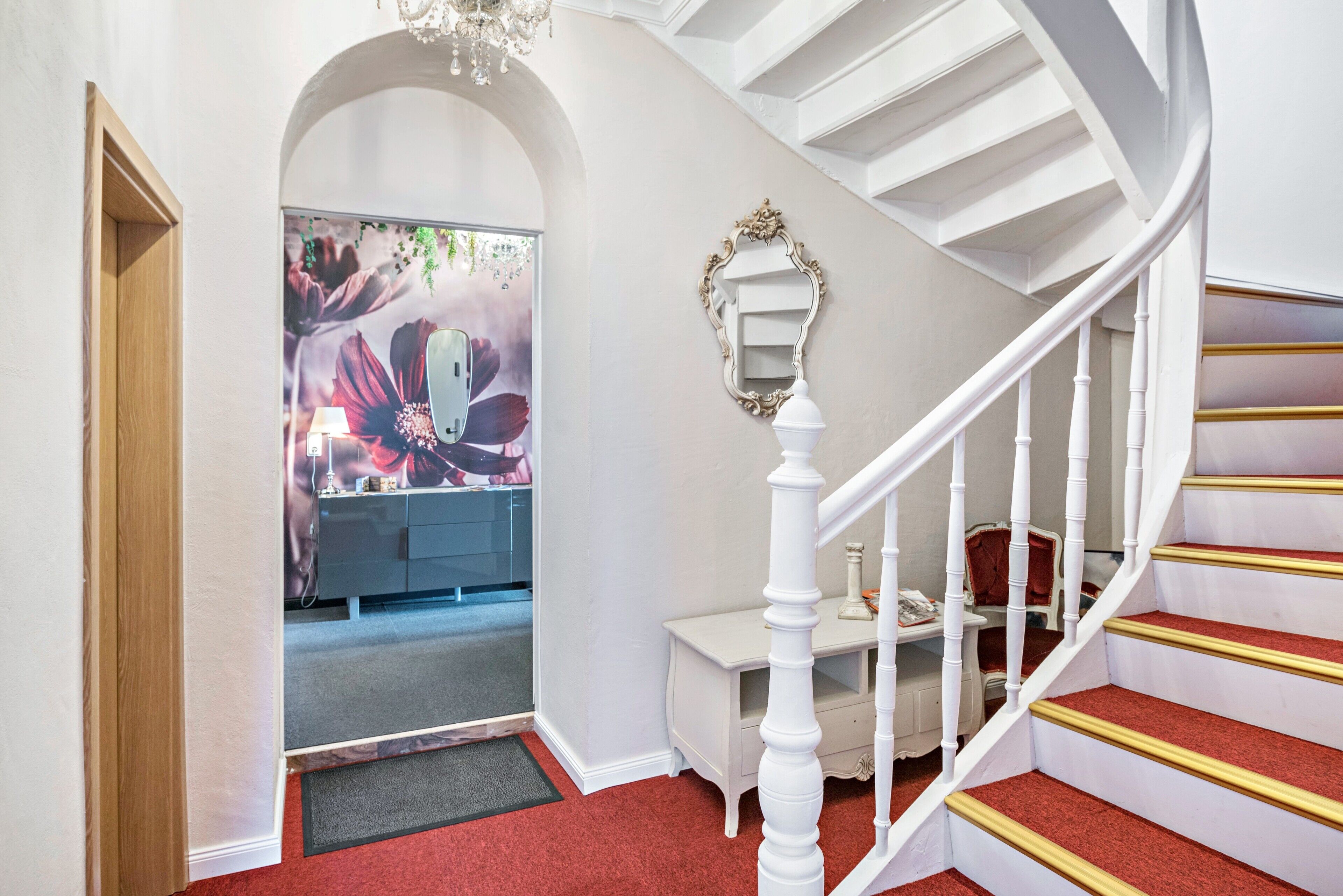 A staircase with white balusters and red carpeted steps, leading to a room with a large floral mural and a white vanity.