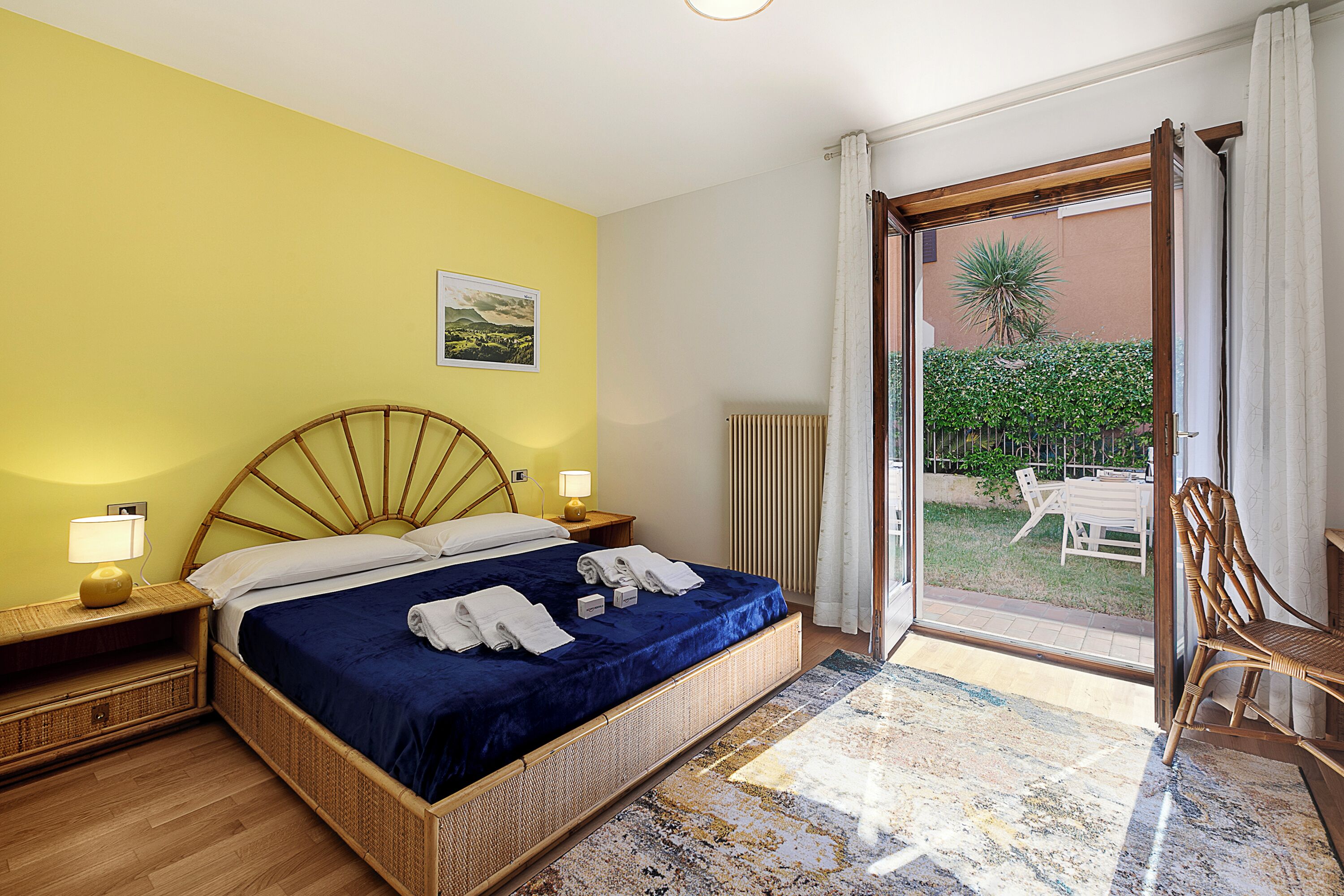 A room with a bed, a nightstand, a lamp, a chair, and a view of the outside.