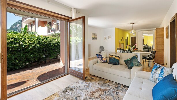 A modern living room with a white sofa, a dining area, and a large sliding glass door leading to a patio. - Il Giardino di Riva - Garden Apartment Near the Lake (Riva del Garda)