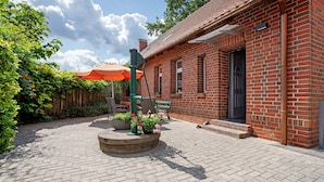 Un bâtiment en briques avec une porte noire, une clôture en bois et une terrasse pavée agrémentée d’un jardinière circulaire et d’un parasol orange.