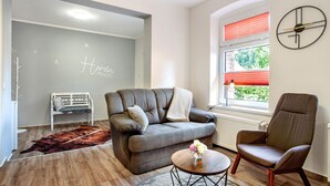 Un salon avec un canapé gris, une table basse en bois, un fauteuil marron, un store à rayures rouges et une horloge murale.